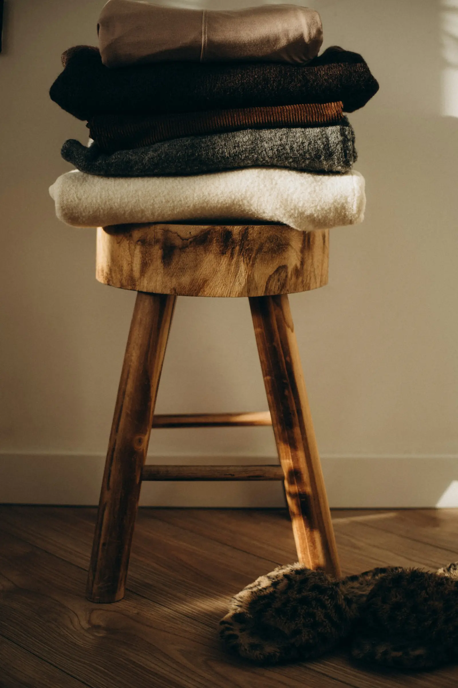 A stack of folded woollen sweaters in brown, black, grey and cream on a wooden stool