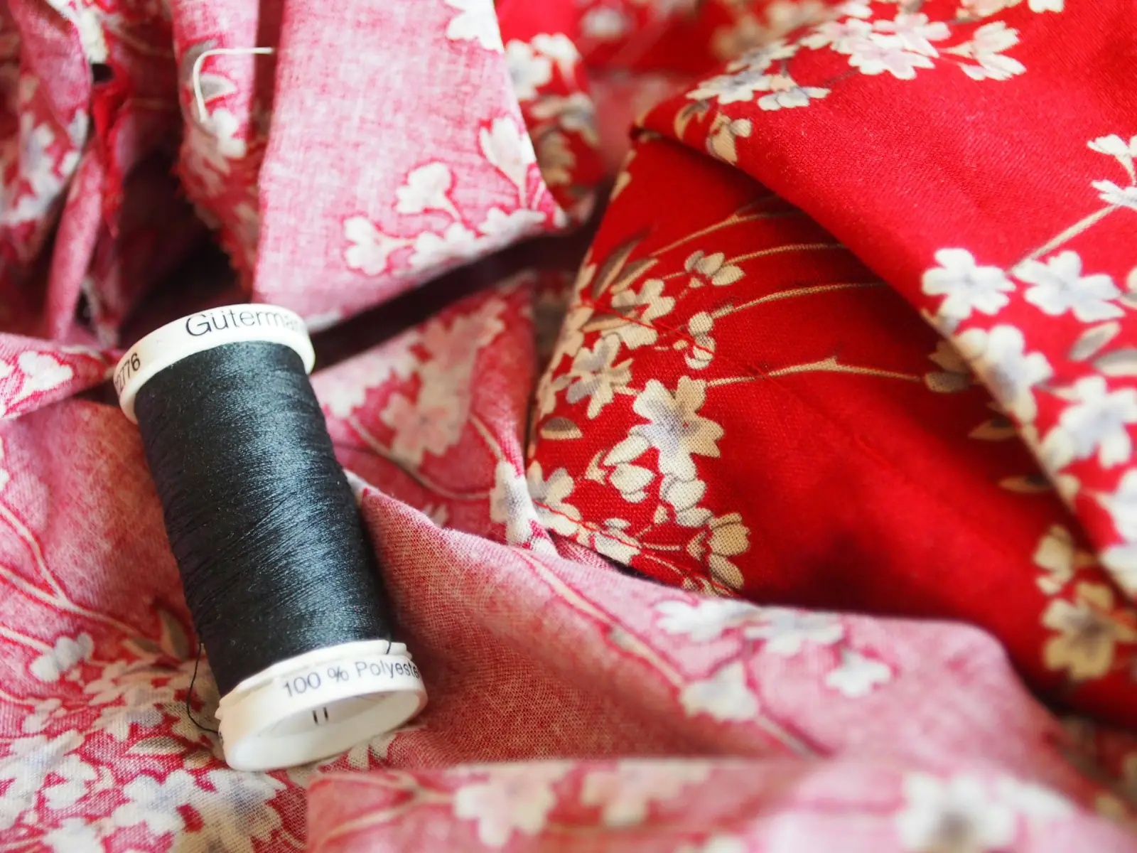 A spool of dark thread resting on folds of red and pink cotton printed with small white flowers