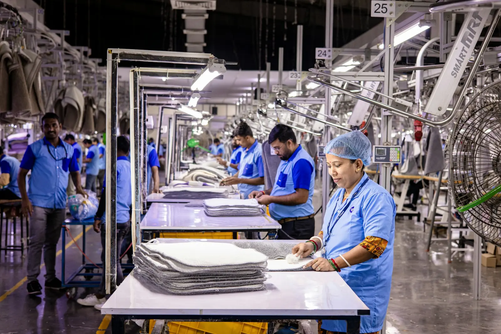 Workers in blue uniforms finishing and stacking garments at long tables on an Indian factory floor