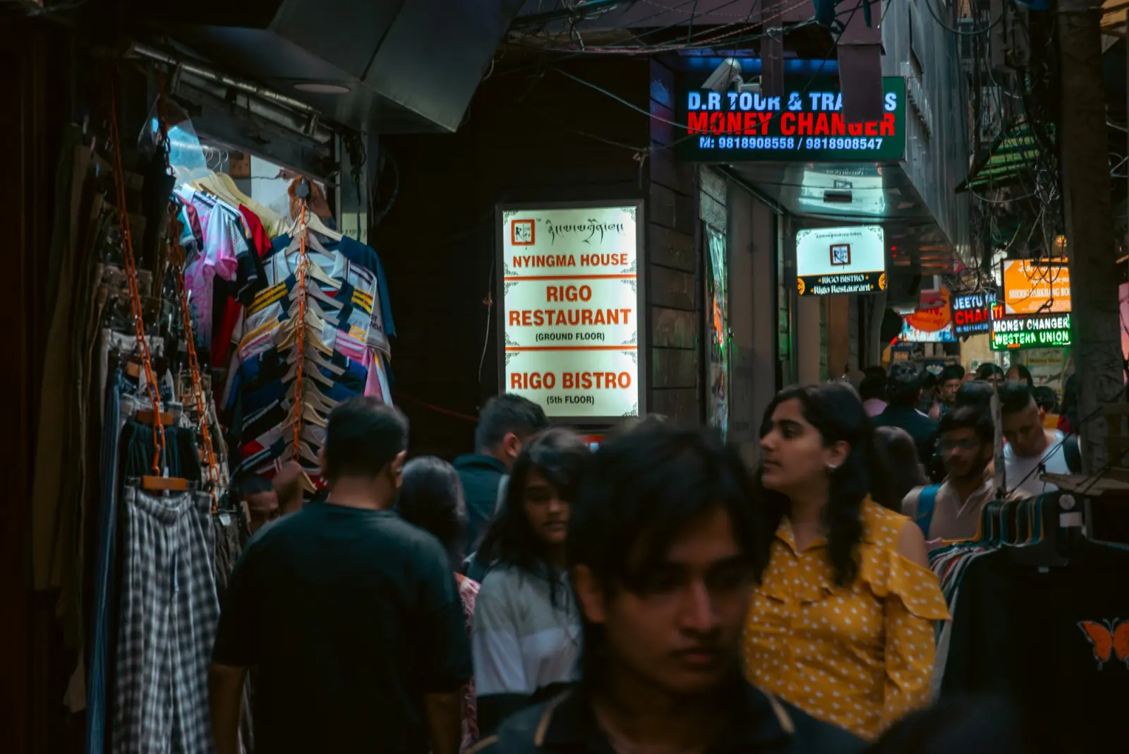 A crowded, narrow Delhi market lane at dusk with clothes hanging from shopfronts and bright signboards overhead