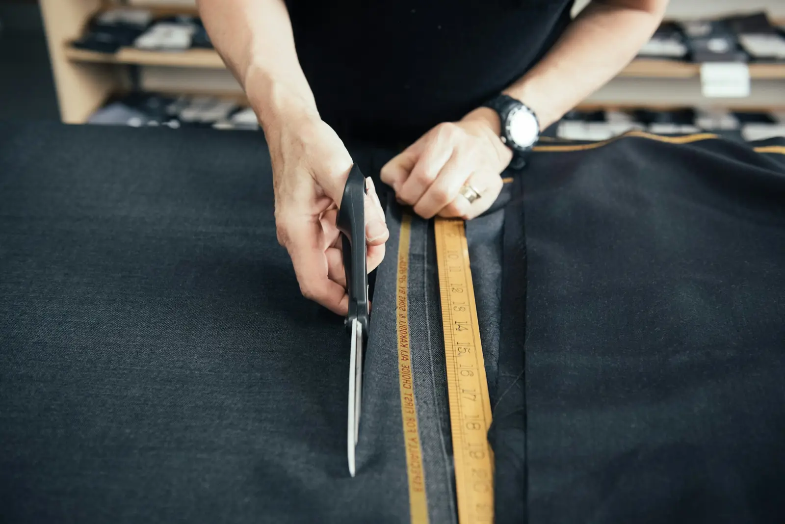 A shopkeeper measuring dark suiting fabric with a wooden ruler and cutting it with large tailoring scissors