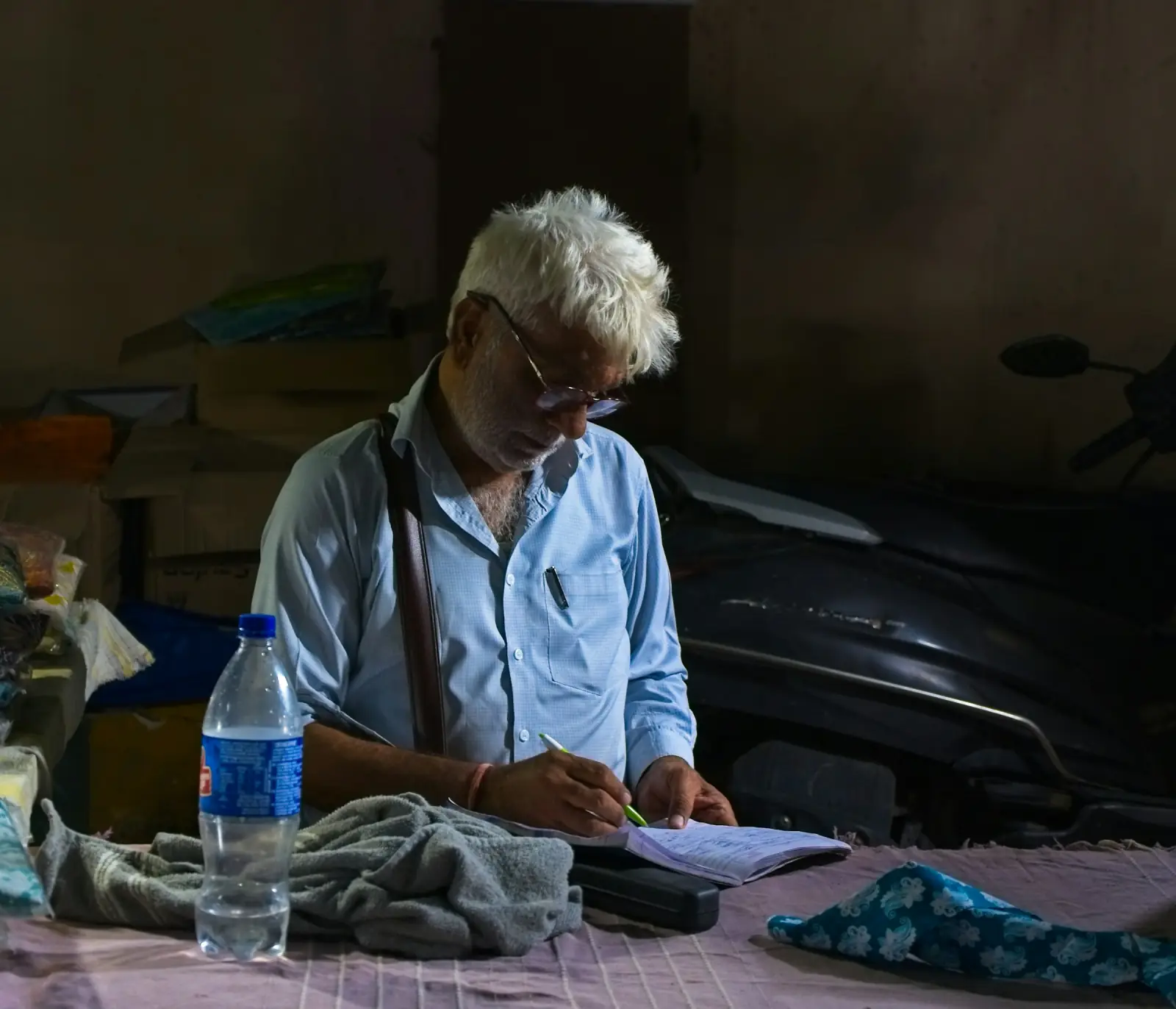 An older shopkeeper in a blue shirt writing entries into a small notebook at a cluttered desk