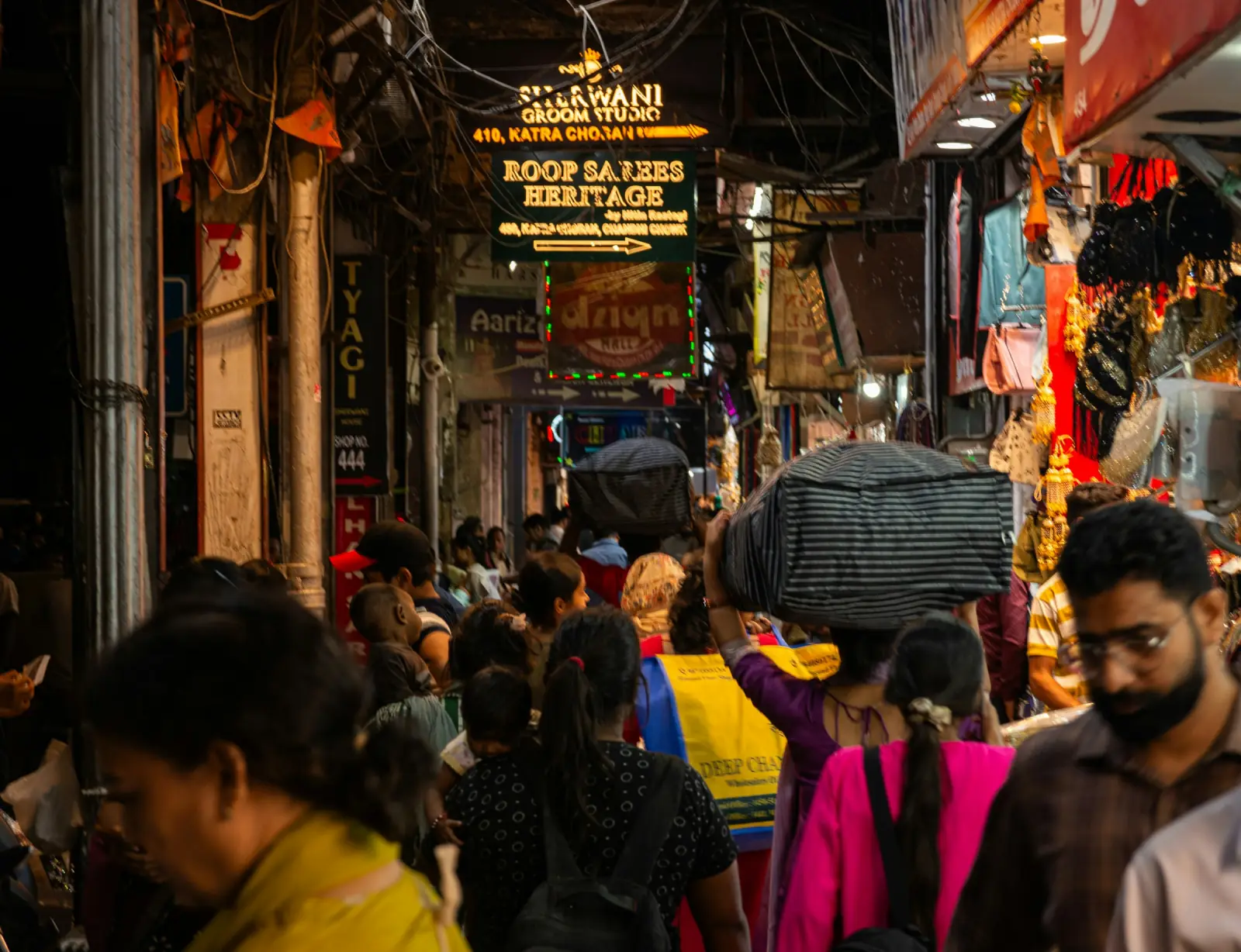 A crowded Indian textile market lane at dusk, lit by saree and fabric shop signboards