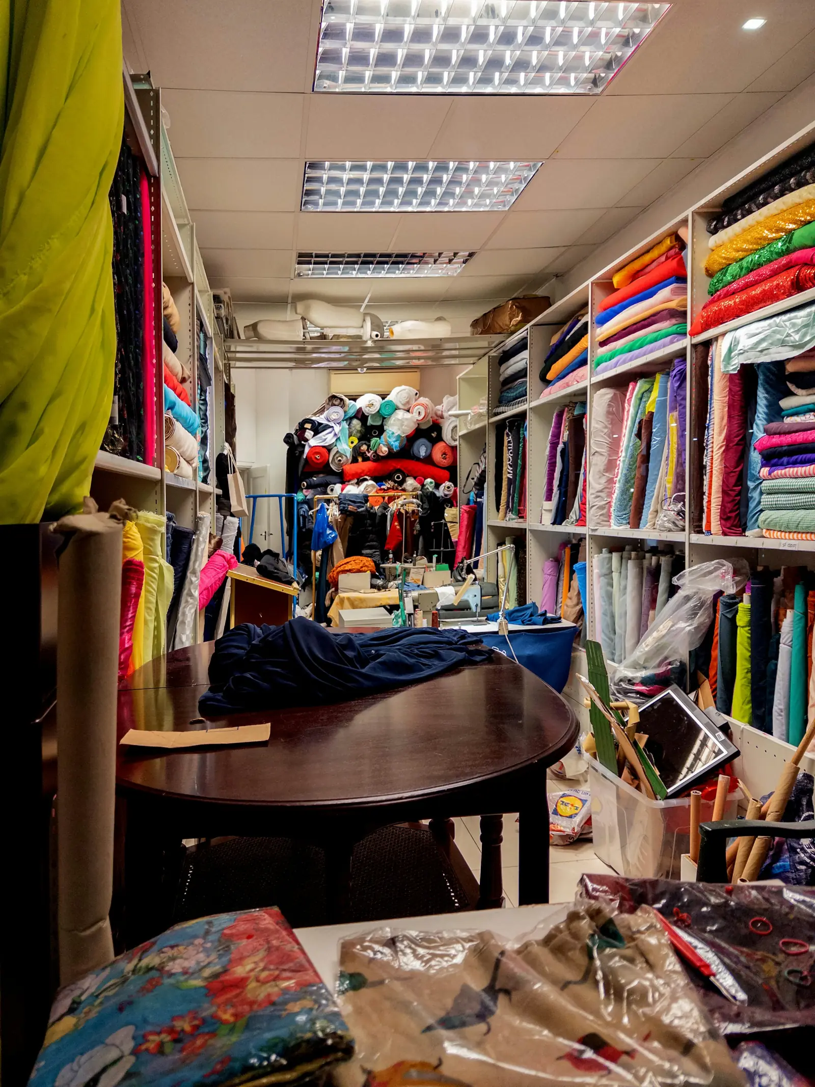 A cramped fabric shop stockroom with shelves of folded cloth, rolls stacked to the ceiling and a cutting table