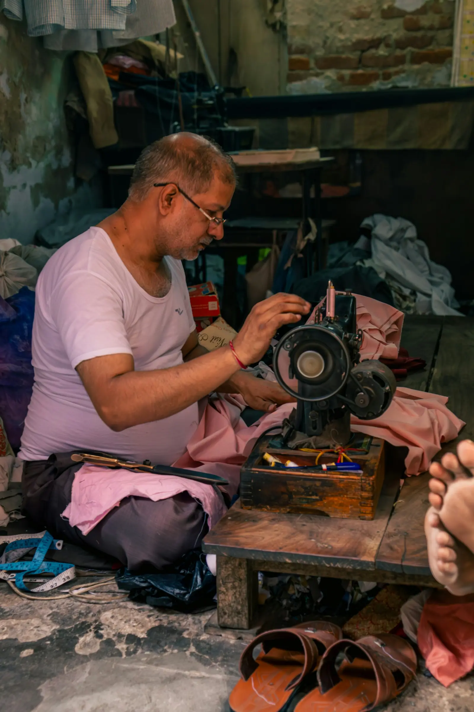 An elderly tailor in a white vest stitching pink fabric on a hand-operated sewing machine in his workshop
