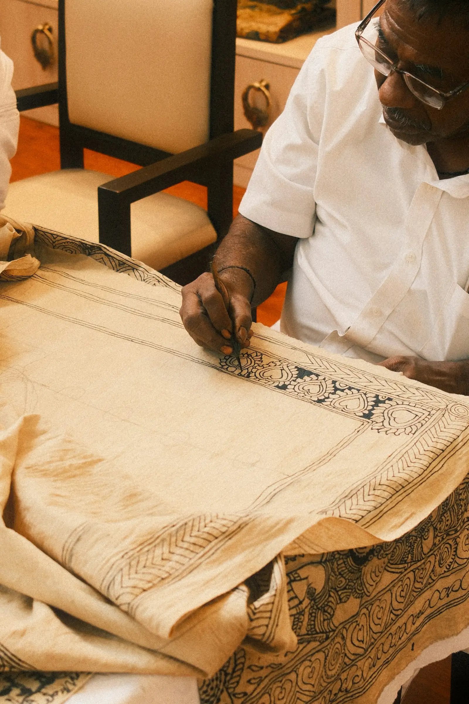An artisan in a white kurta hand-painting a detailed pattern onto a length of pale cotton fabric