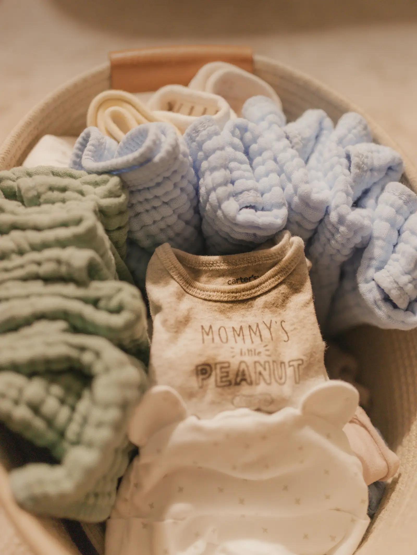 A woven basket filled with soft folded muslin baby swaddles and baby clothes in pastel colours