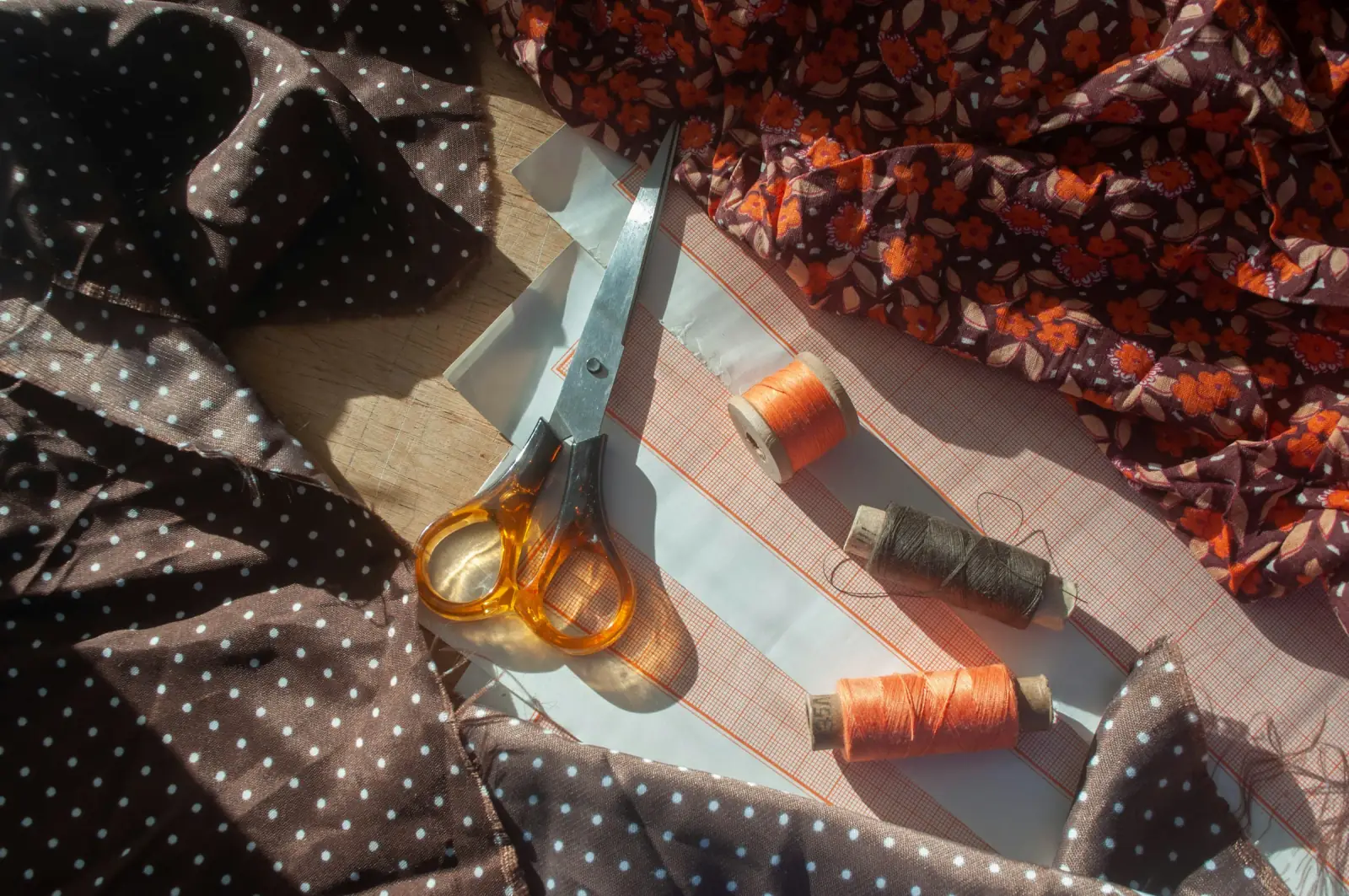Scissors, orange thread spools and a paper pattern lying on dark polka-dot and floral printed fabrics