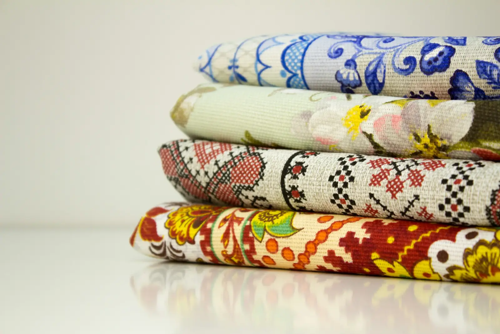 A stack of folded patterned fabrics including blue and white tiles, cross-stitch style and bold floral prints