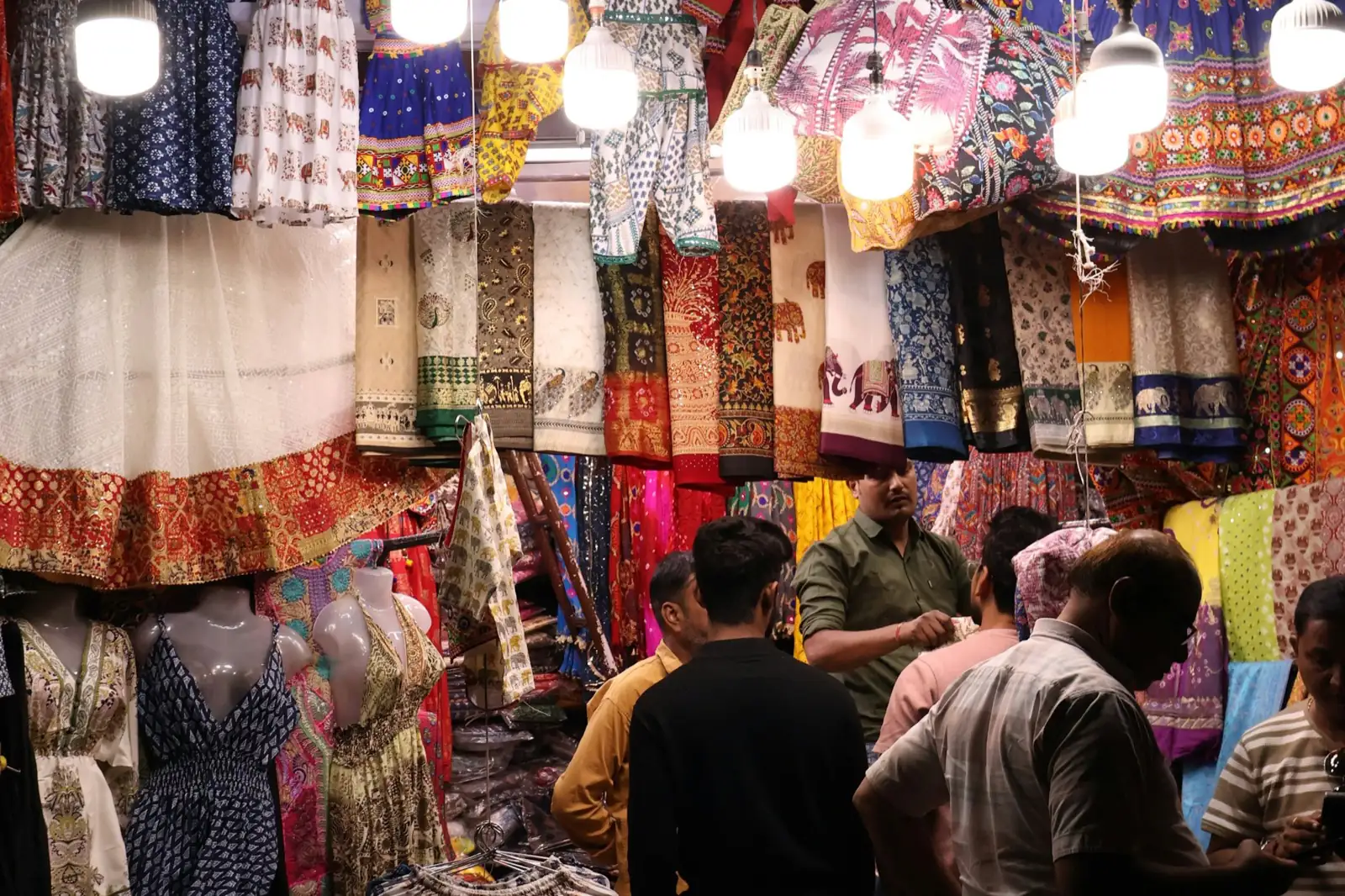 Customers browsing brightly lit racks of clothing and fabric at an Indian market stall