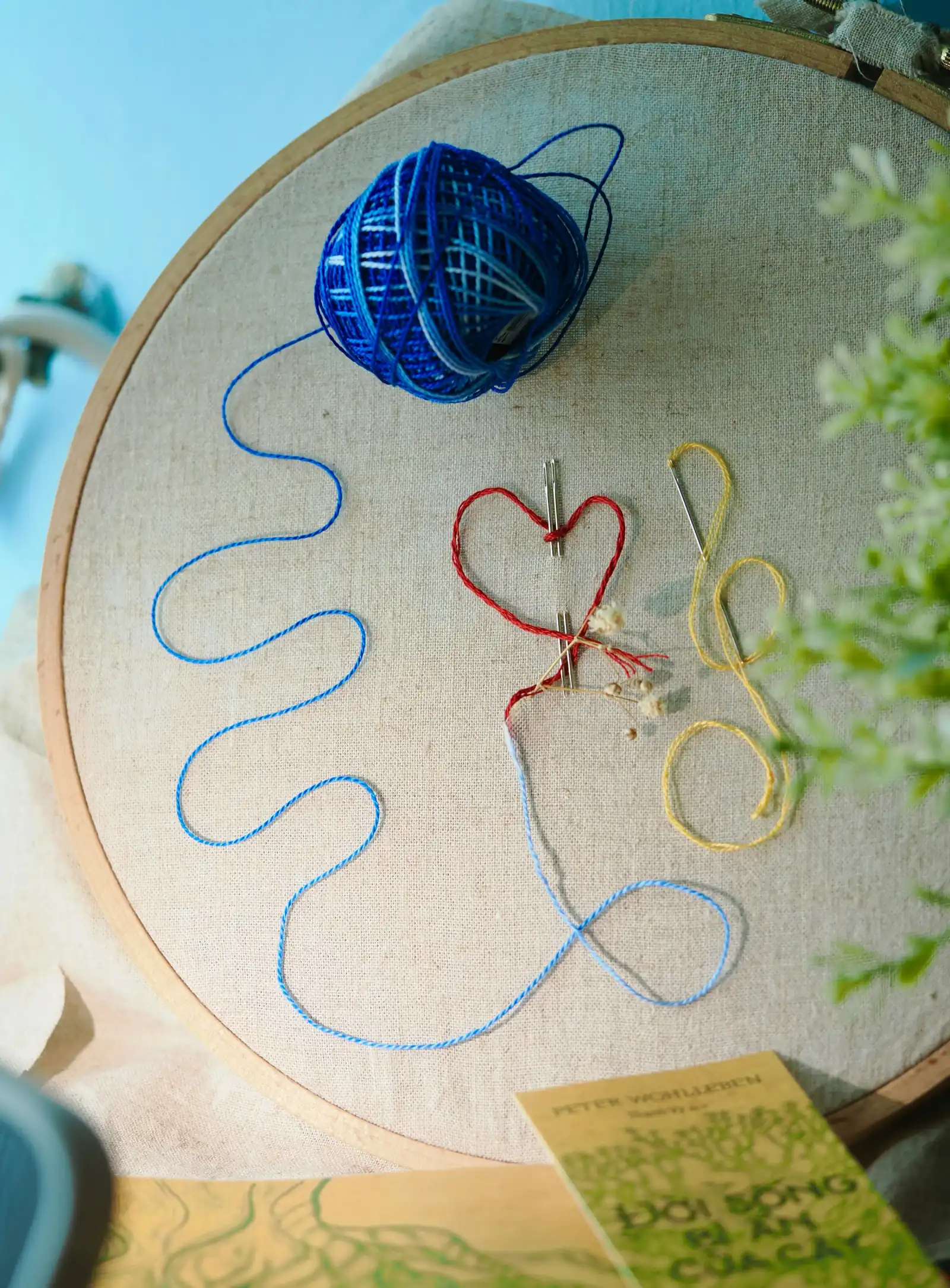 An embroidery hoop with a stitched red heart and a ball of blue yarn with a needle and thread