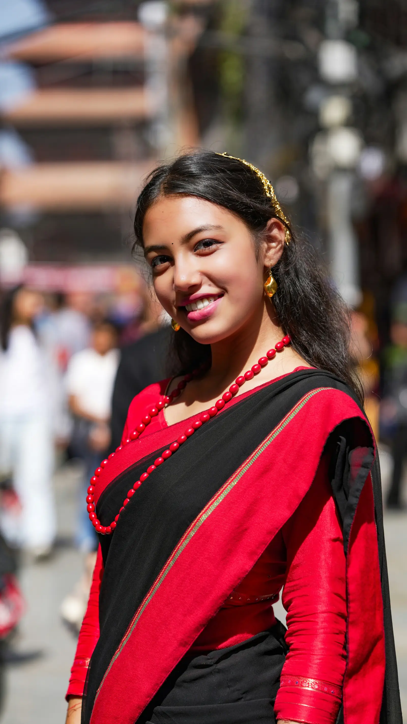A smiling young woman in a red and black sari with gold jewellery standing on a busy street